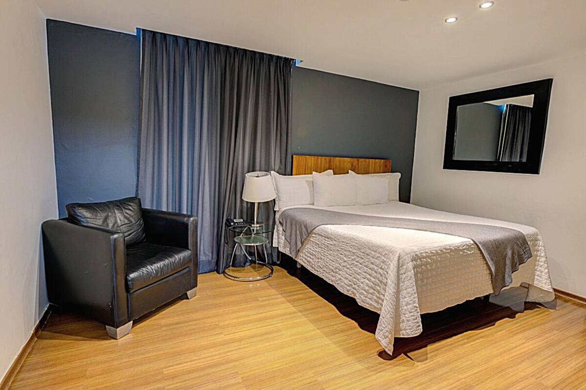 A hotel room with a bed, a chair, a bedside table with a lamp, and a mirror on the wall.