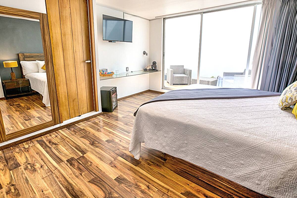 A hotel room with a large bed, wooden flooring, a TV, a balcony with a chair, and a mirror.