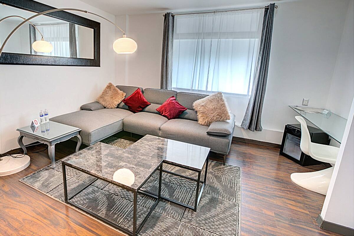 A modern living room with a sectional sofa, a glass coffee table, and a dining table. There is a large window with curtains, a mirror on the wall, and a small side table with water bottles.