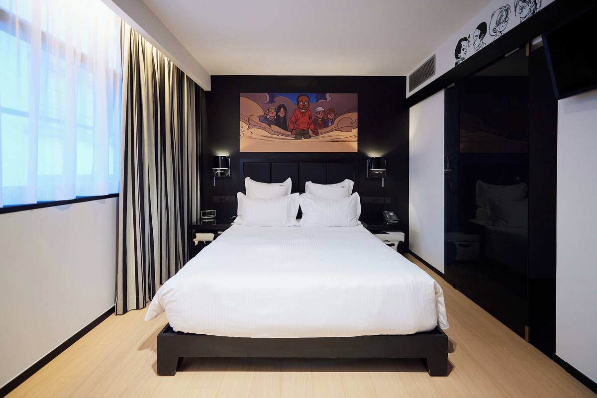 A modern hotel room with a large bed, a headboard with a mural, bedside lamps, and a wardrobe.