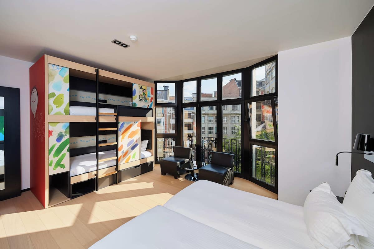 A modern hotel room with a large bed, a built-in bookshelf, a desk, and a view of the city through large windows.