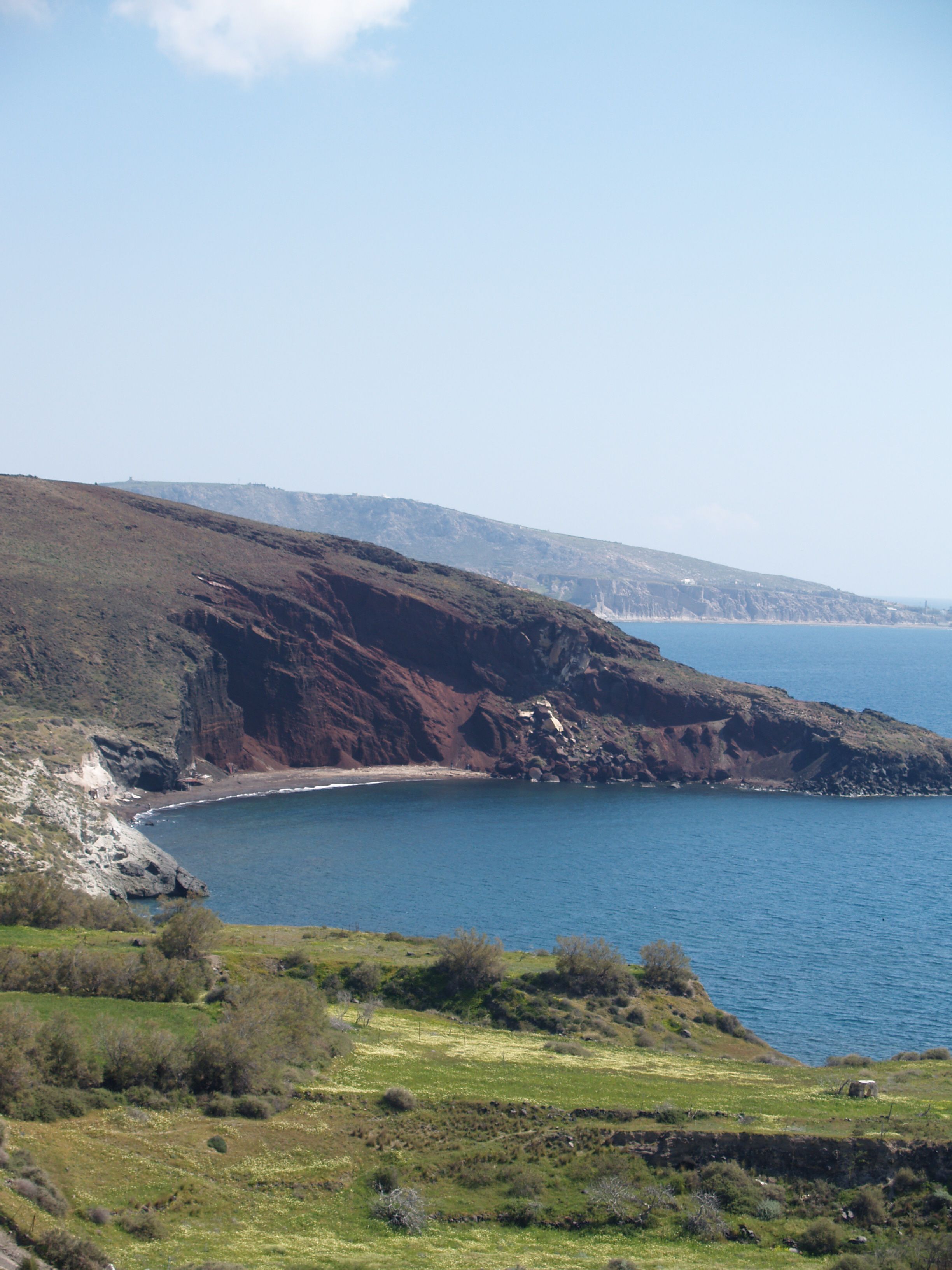 A coastal landscape with a rocky shoreline, a clear blue sea, and a hilly terrain with sparse vegetation.