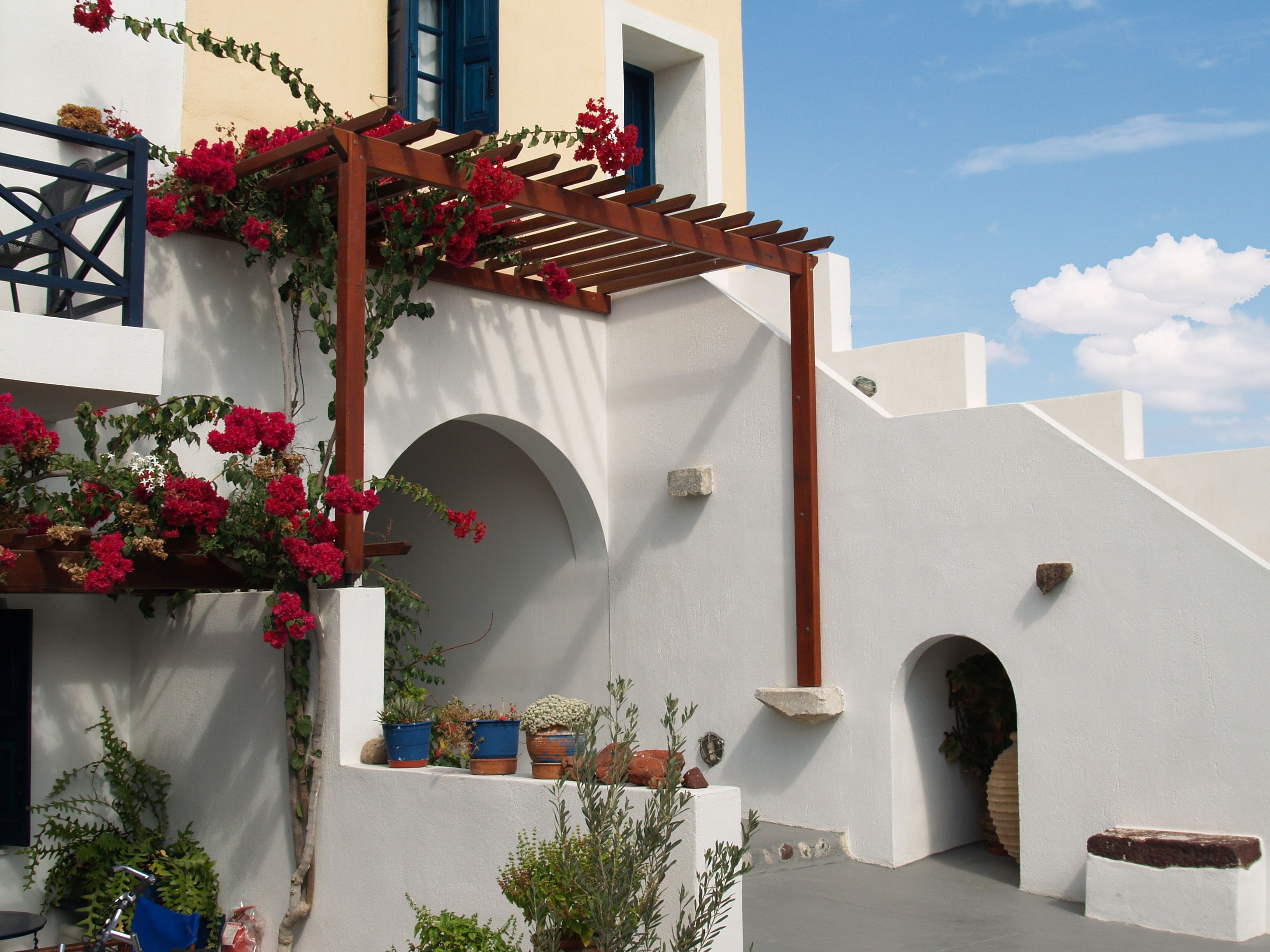 A building with white walls, a wooden pergola with red flowers, and multiple potted plants.