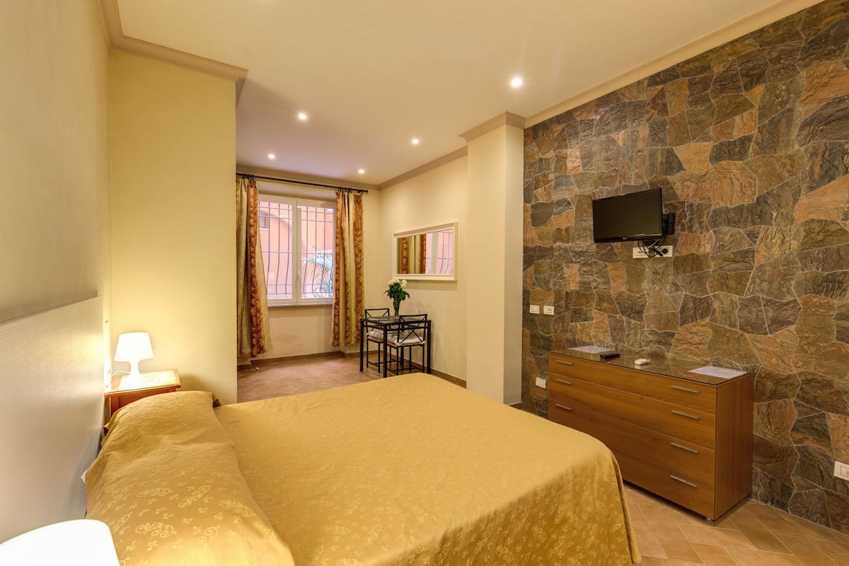 A hotel room with a bed, a dresser, a TV mounted on a stone wall, a small table with chairs, and a window with curtains.
