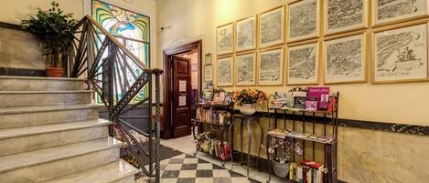 Um saguão de hotel com piso xadrez, escada com corrimão de metal, parede com mapas emoldurados e balcão de recepção com livros e uma planta em vaso.