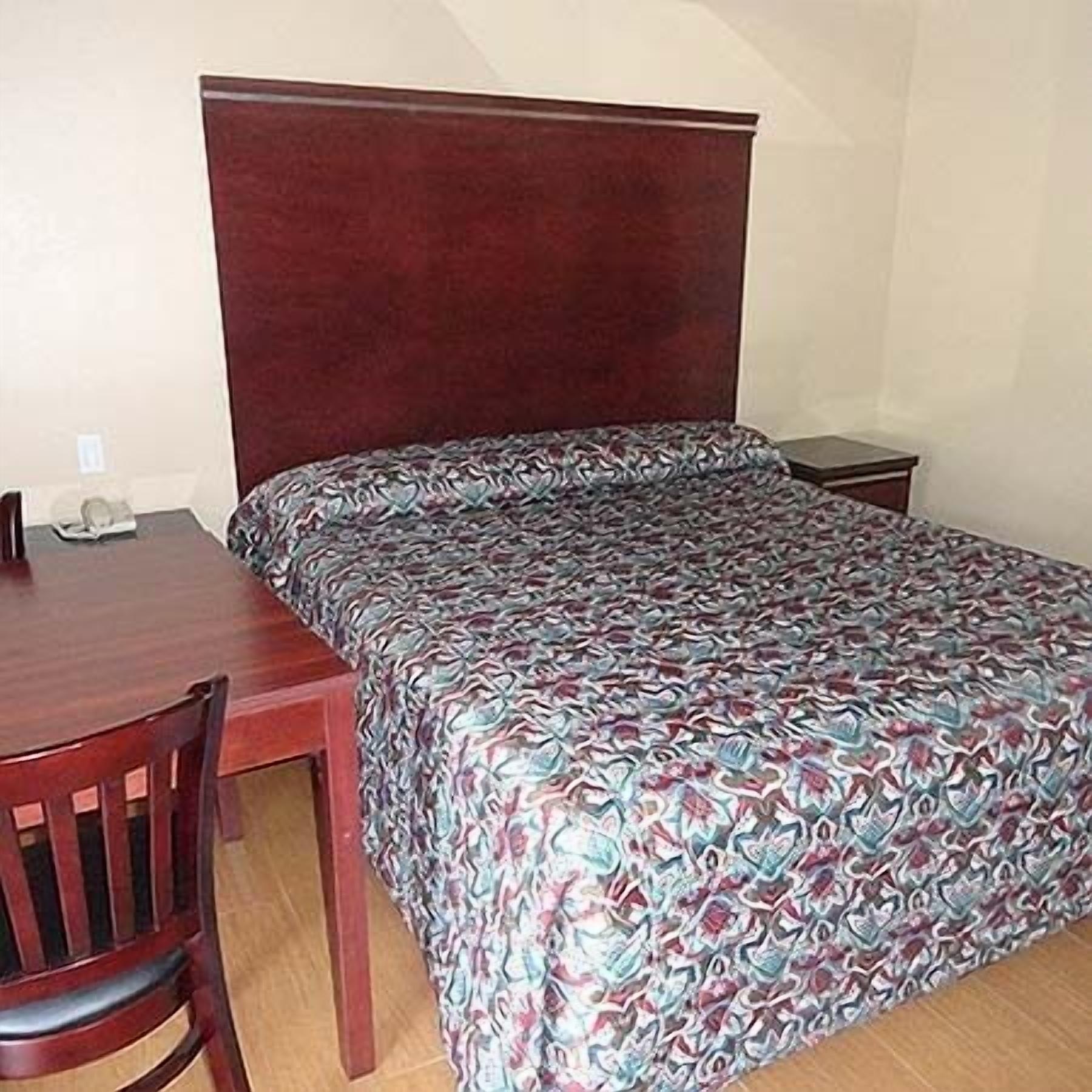 A single bed with a patterned bedspread, a wooden nightstand, a wooden desk with a chair, and a small table with a telephone.