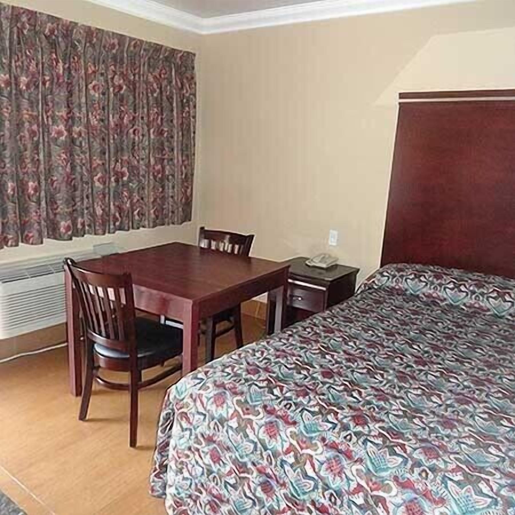 A hotel room with a wooden dining table, two chairs, a bed with a patterned bedspread, a nightstand, and a window with floral curtains.