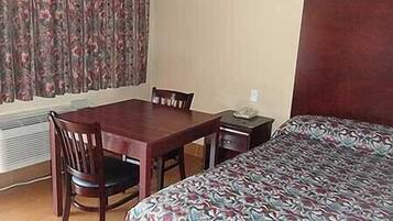 A hotel room with a wooden dining table, two chairs, a bed with a patterned bedspread, a nightstand, and a window with floral curtains.