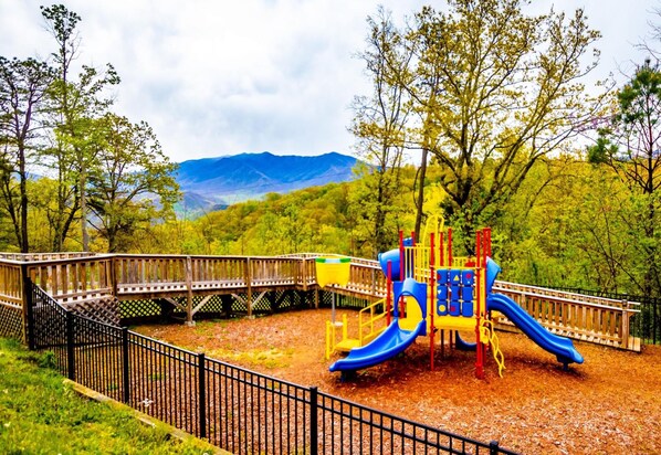 A playground with a slide and climbing structures, surrounded by a wooden fence and trees, with mountains in the background. - Laurel Point Resort (Gatlinburg)