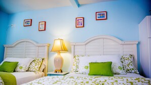 Habitación de hotel con dos camas, cada una con cabecera blanca, ropa de cama blanca y verde con motivos florales, y una mesita de noche con lámpara.