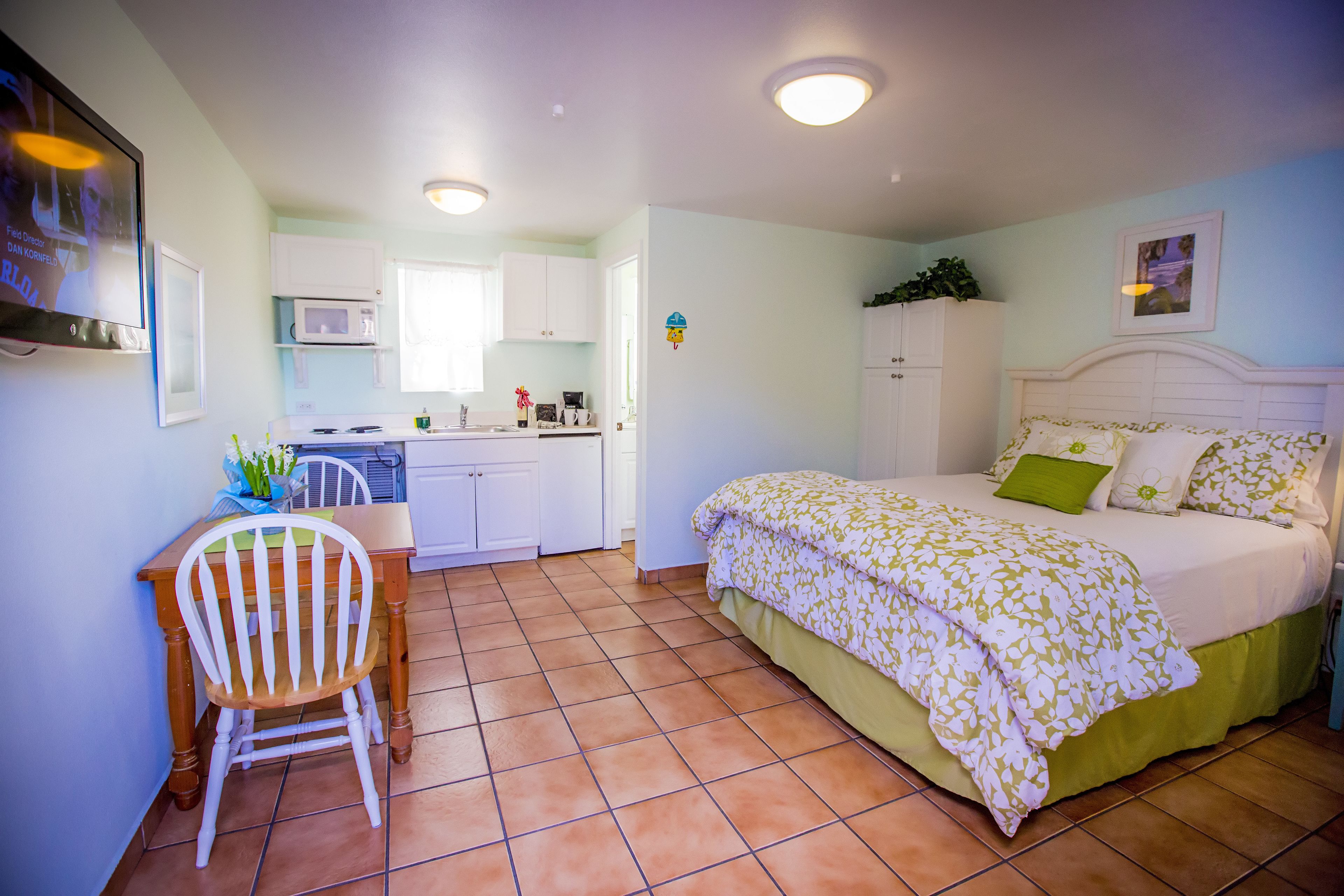 A bedroom with a bed, a desk, and a chair. There is a kitchen area with cabinets and a microwave.