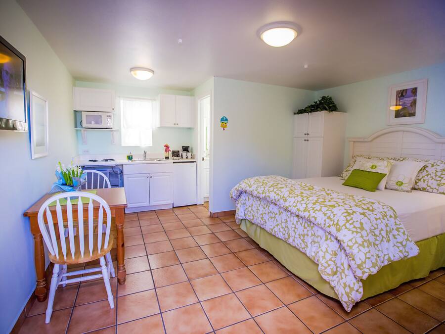 A bedroom with a bed, a desk, and a chair. There is a kitchen area with cabinets and a microwave.