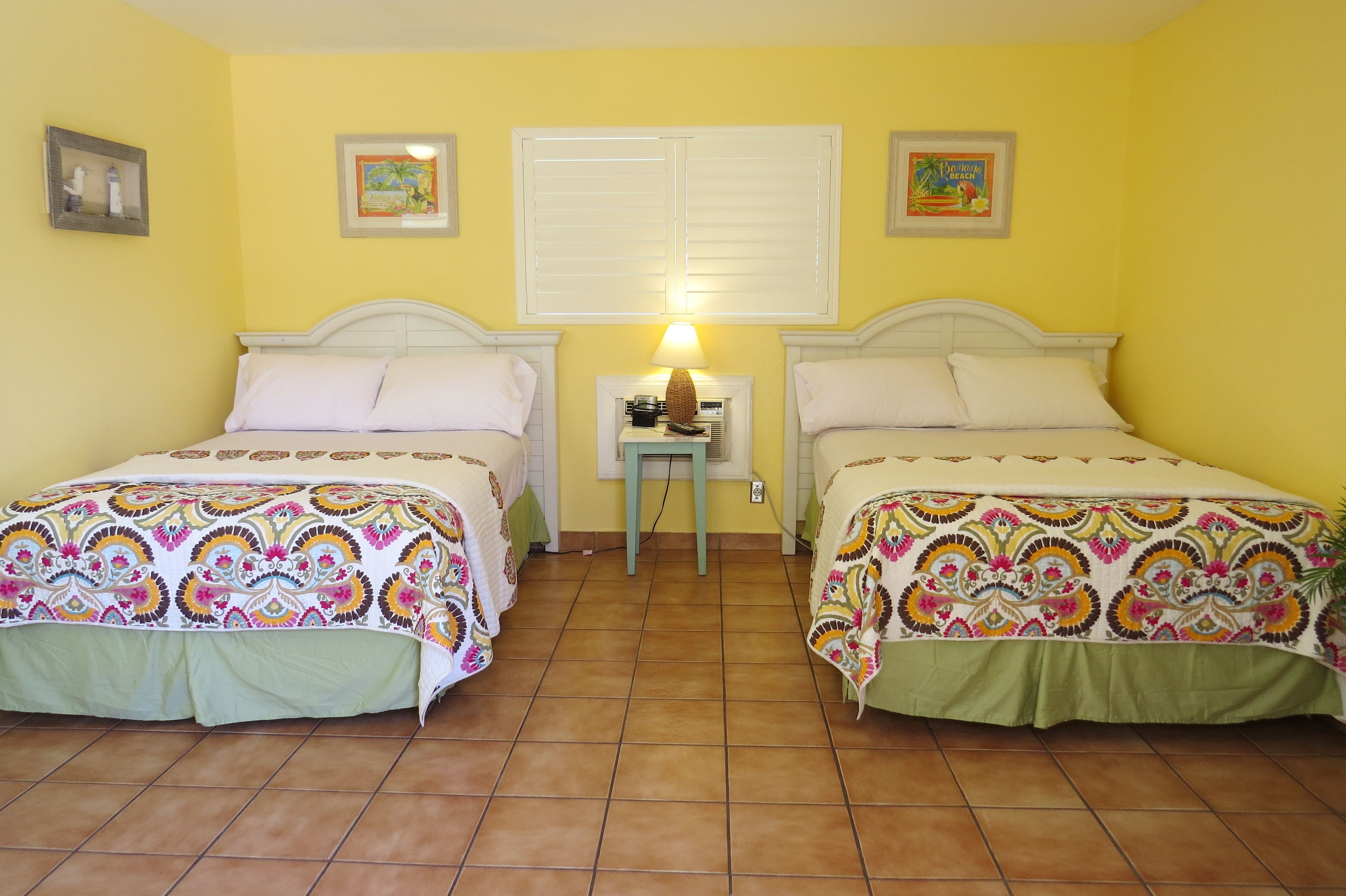 A room with two beds, a small table with a lamp, and a window with shutters.