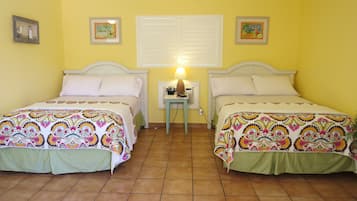 A room with two beds, a small table with a lamp, and a window with shutters.