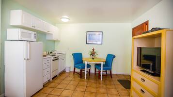 A compact kitchen with white appliances, a dining area with blue chairs, and a TV on a wooden cabinet.