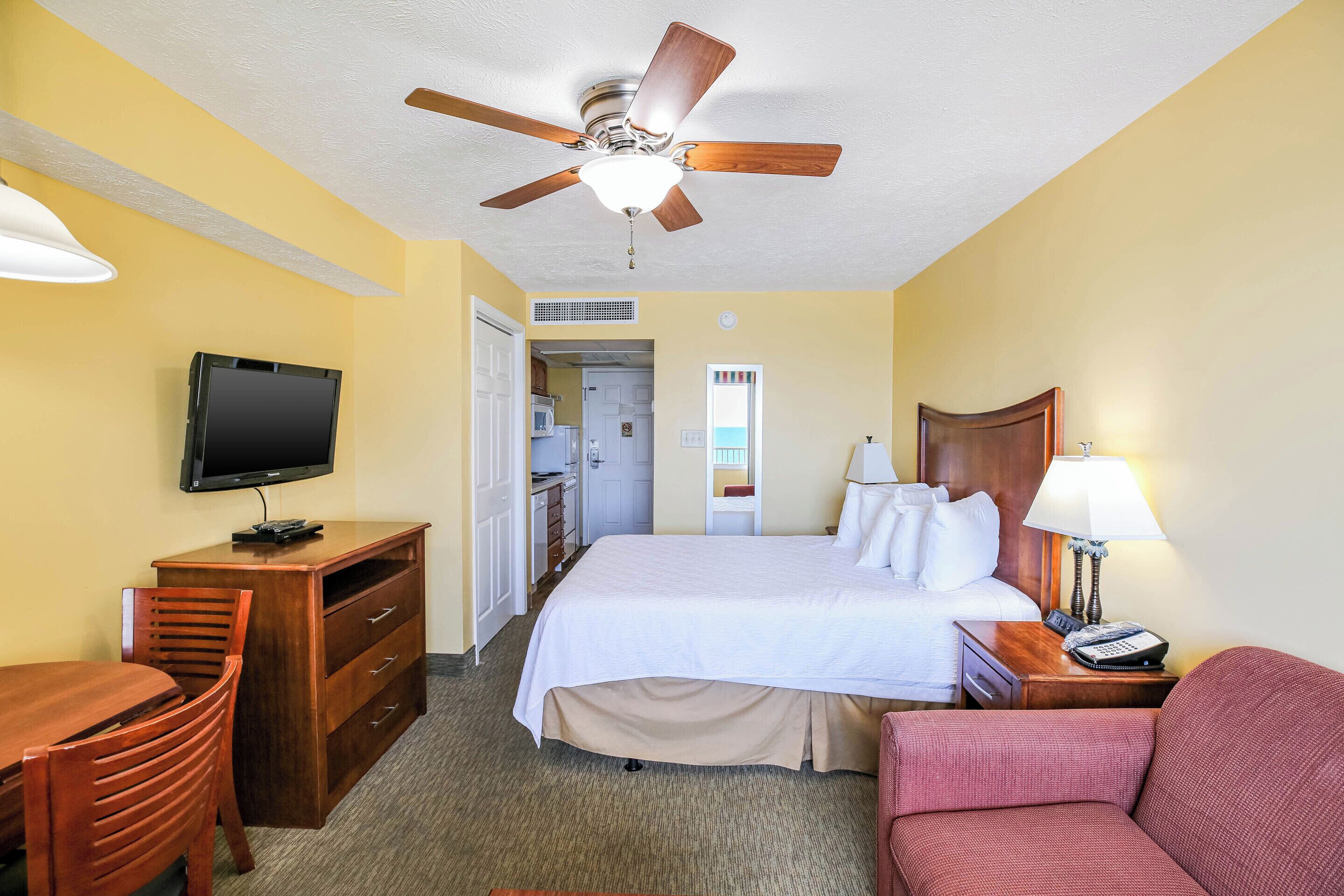Une chambre d’hôtel comprenant un lit, un bureau avec une télévision, une chaise, un canapé et un ventilateur de plafond.
