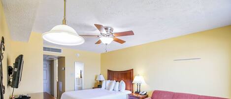 Una habitación de hotel con una cama, un sofá rojo, una mesa de madera, un ventilador de techo y un televisor sobre un mueble.
