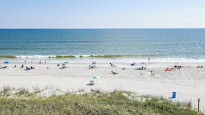 Ein Strand mit Menschen, die unter Sonnenschirmen entspannen, ein sandiger Uferstreifen und das Meer im Hintergrund.