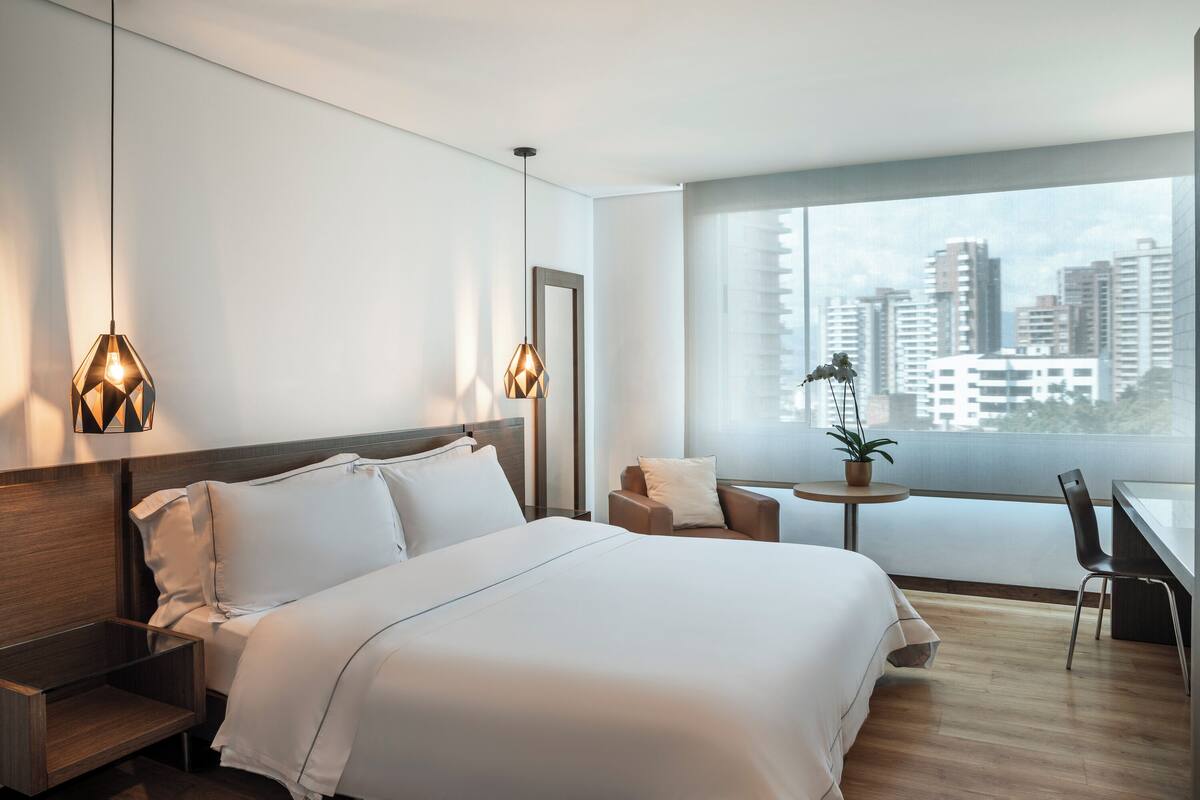 A modern hotel room with a large bed, a desk, a chair, and a view of the city skyline through large windows.