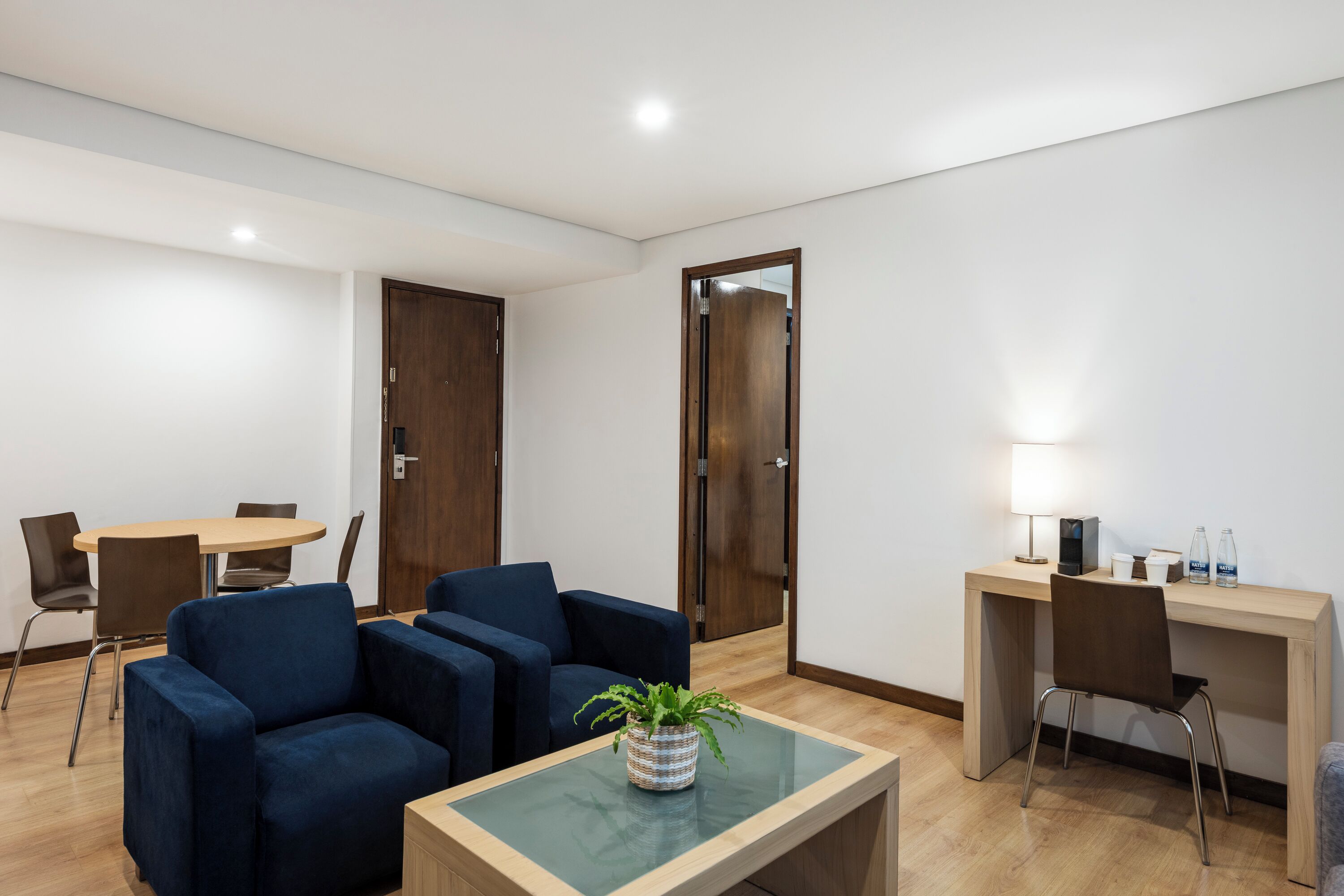 A modern room with a wooden desk, two blue armchairs, a small table with a plant, and a dining table with chairs.