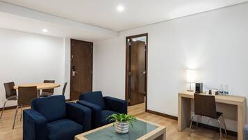 A modern room with a wooden desk, two blue armchairs, a small table with a plant, and a dining table with chairs.
