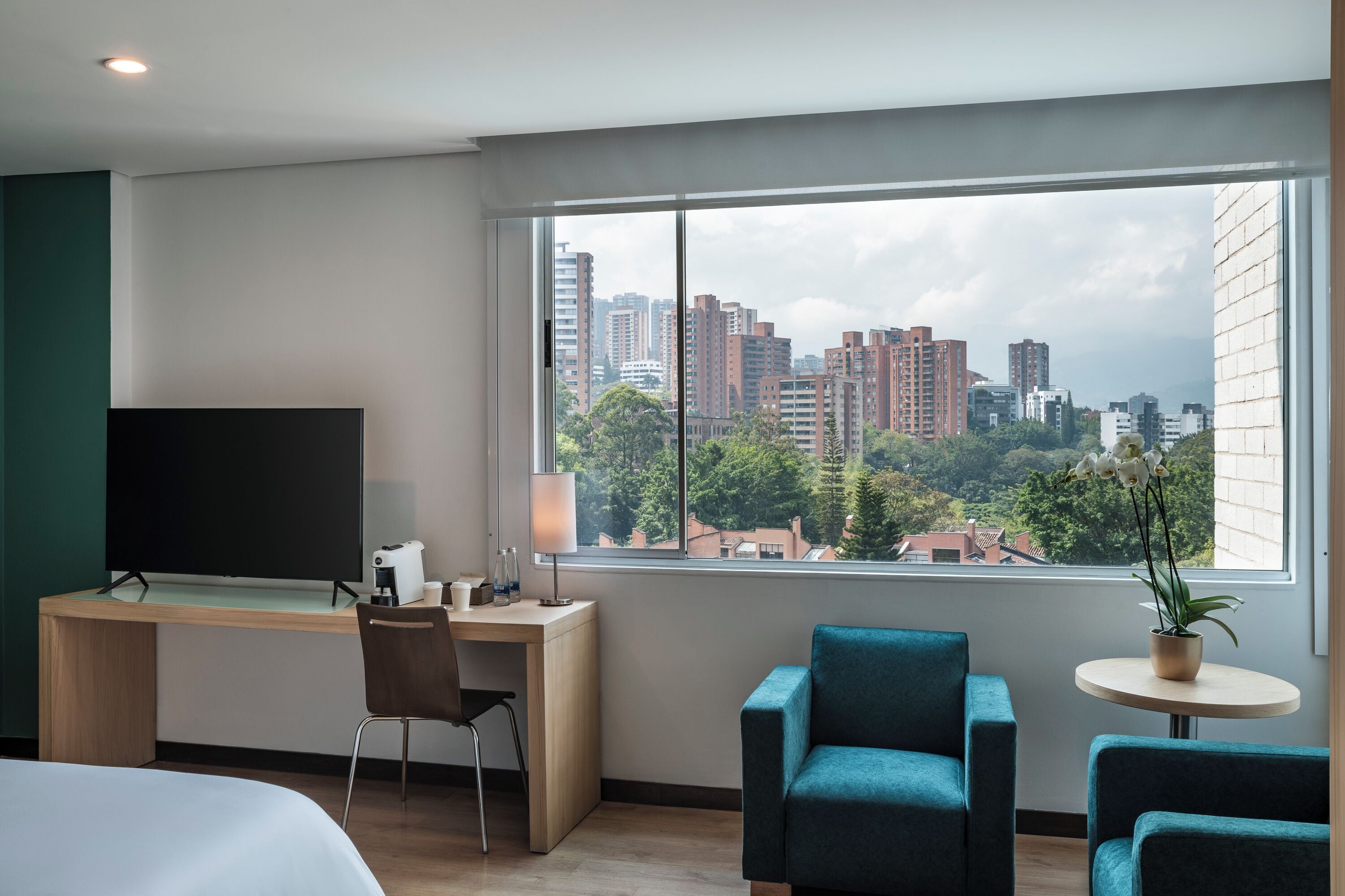 A hotel room with a large window, a desk with a flat-screen TV, a chair, a bed, and two armchairs.