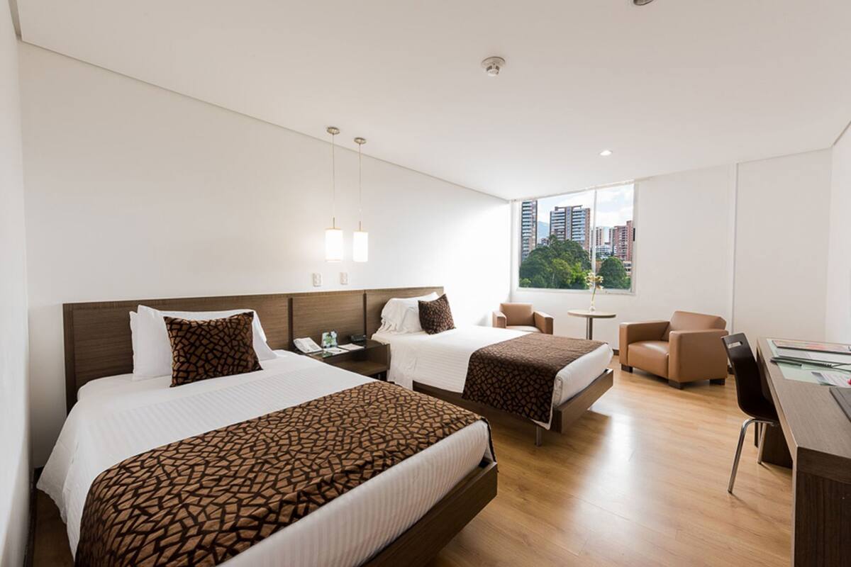 A hotel room with two beds, a desk, a chair, and a window with a city view.