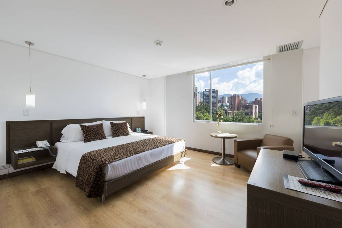 A hotel room with a bed, bedside table, TV, desk, chair, and a window with a city view.