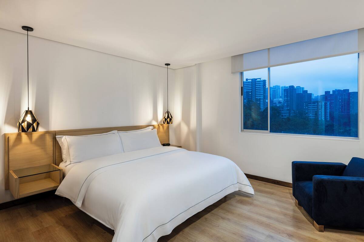 A modern hotel room with a large bed, bedside tables, a chair, and a city view.