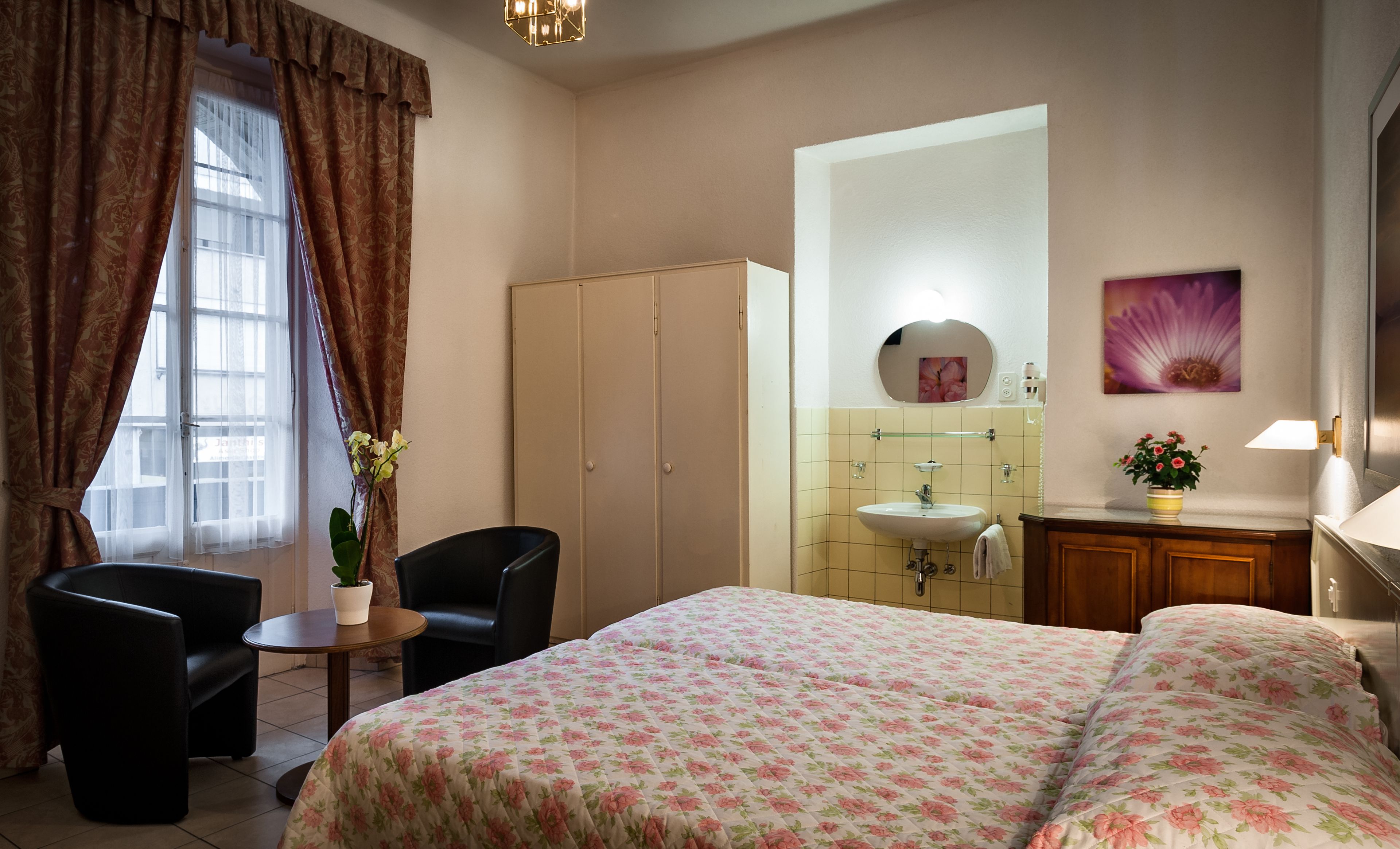 Une chambre d’hôtel comprenant un lit, une chaise, une petite table avec une plante, une armoire, une salle de bain avec un lavabo et un miroir, et un tableau accroché au mur.