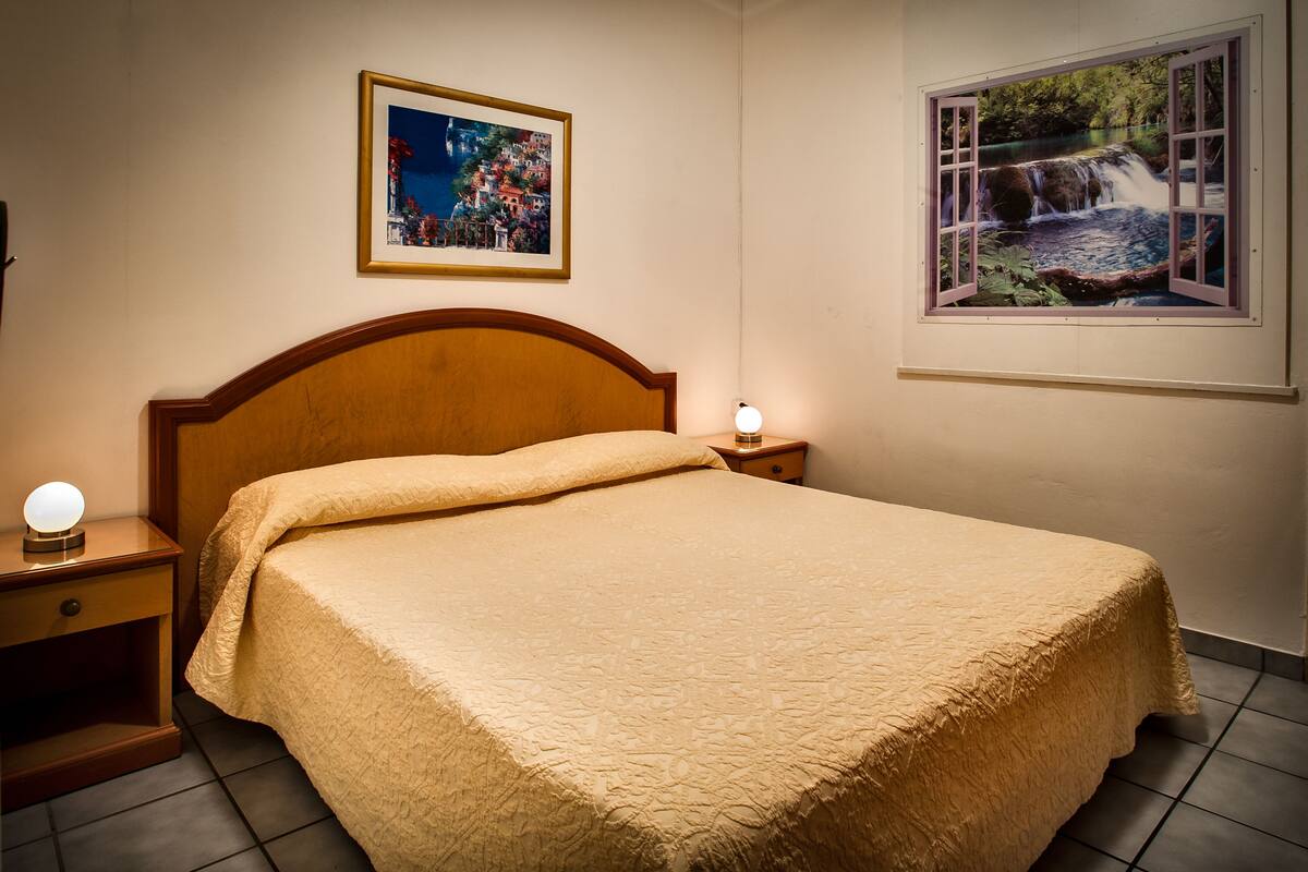 A neatly made bed with a headboard, a bedside lamp, a framed picture on the wall, and a window with a view of a waterfall.