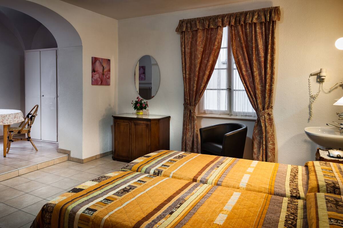 A hotel room with a bed, a desk with a chair, a dresser, a mirror, a window with curtains, and a bathroom with a sink and mirror.