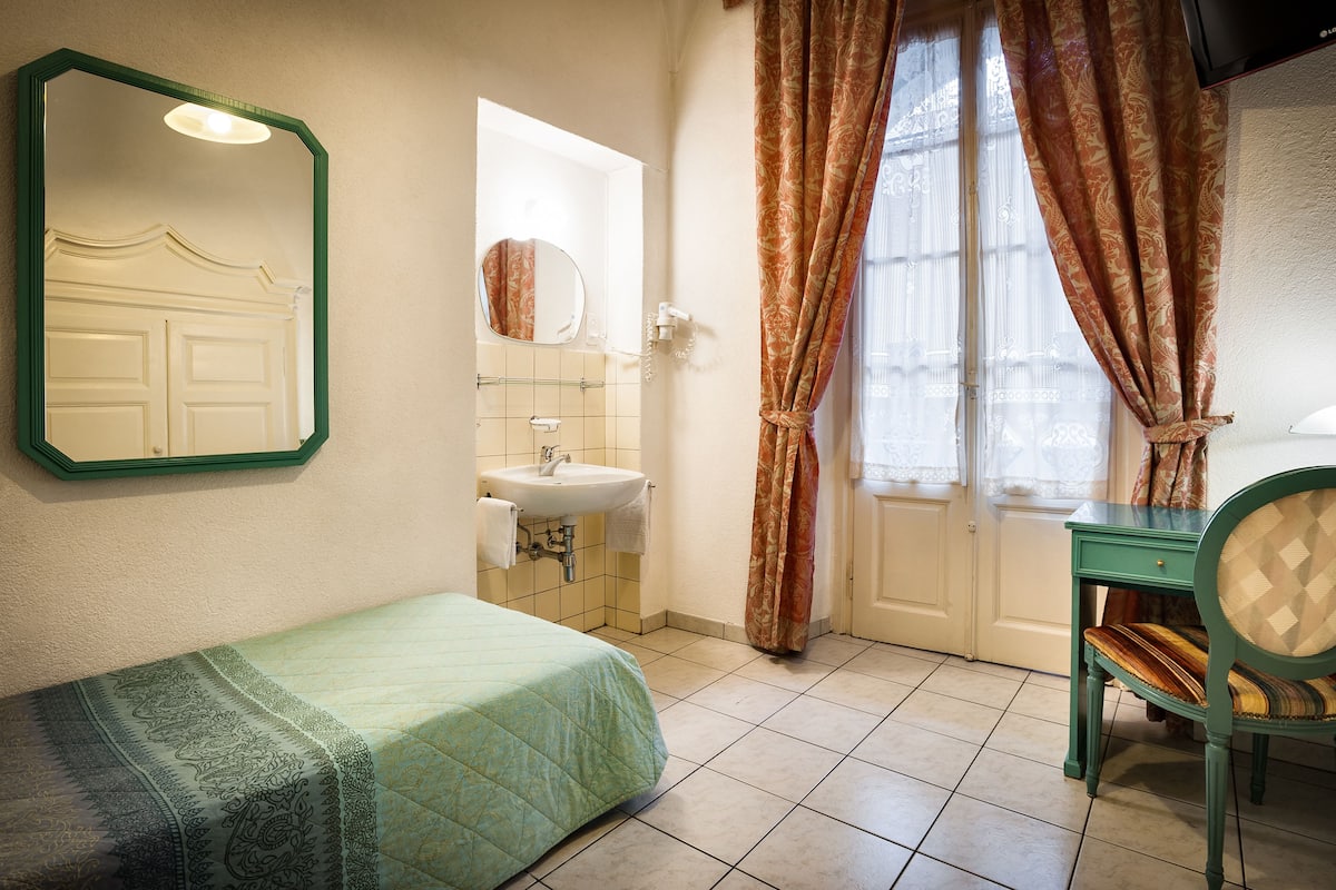 A room with a bed, a green chair, a mirror, a sink, and a window with curtains.