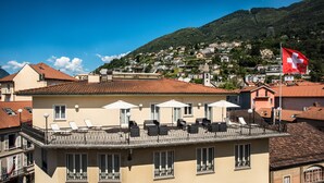A hotel with a terrace featuring outdoor seating and umbrellas, a red flag, and a mountainous backdrop. - EasyRooms dell'Angelo (Locarno)