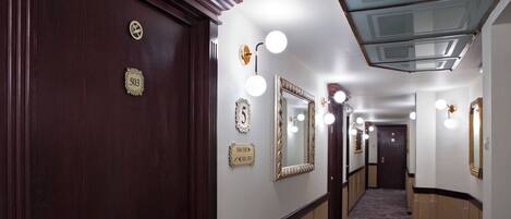 A hotel hallway with a wooden door, numbered plaques, mirrors, and wall sconces.