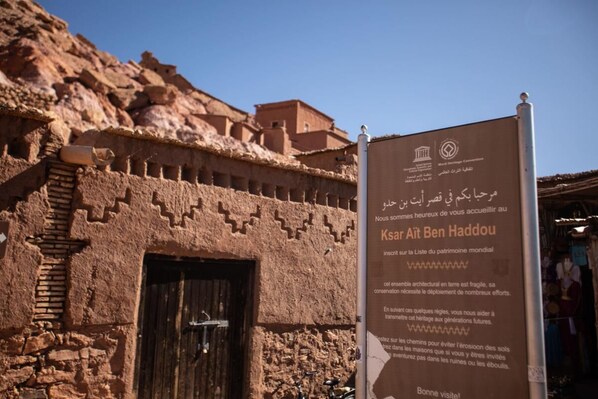 A sign for Ksar Ait Ben Haddou, a UNESCO World Heritage site, with a traditional mud-brick building in the background. - Hotel Le Fint (Ouarzazate)