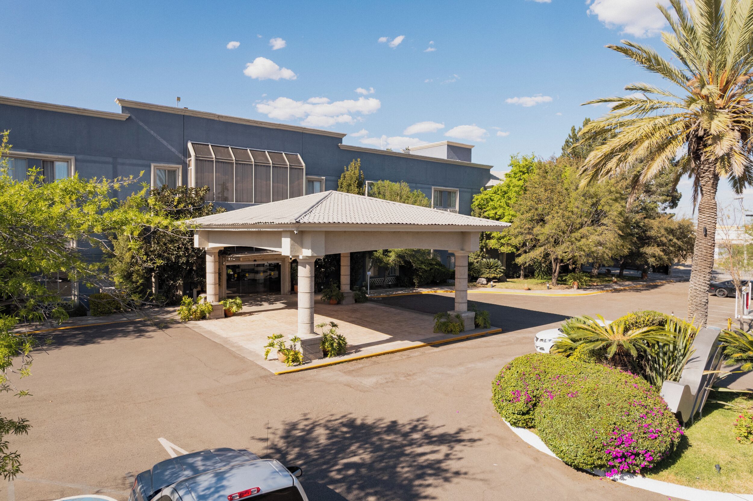 Ein Hotel mit Eingangsbereich, überdachter Terrasse, Palmen und Parkplatz.