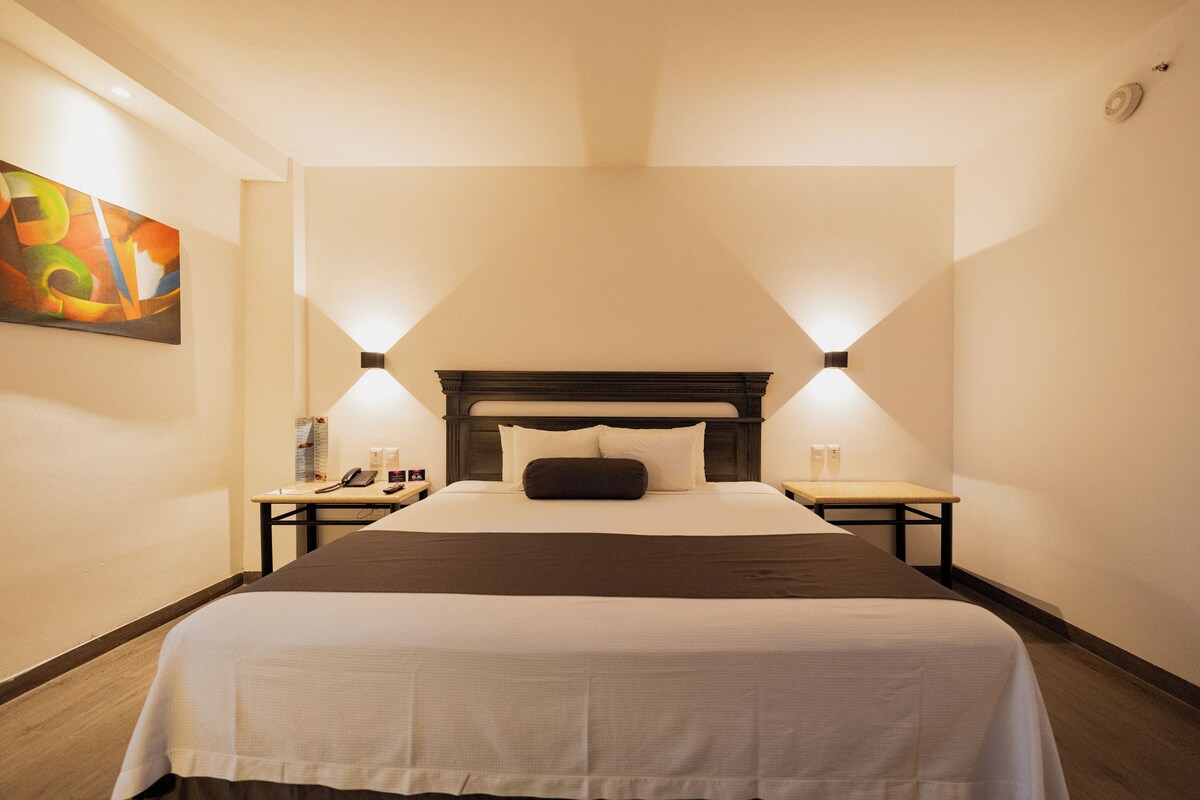 A hotel room with a large bed, two bedside tables, a nightstand, a wall-mounted painting, and a ceiling light fixture.