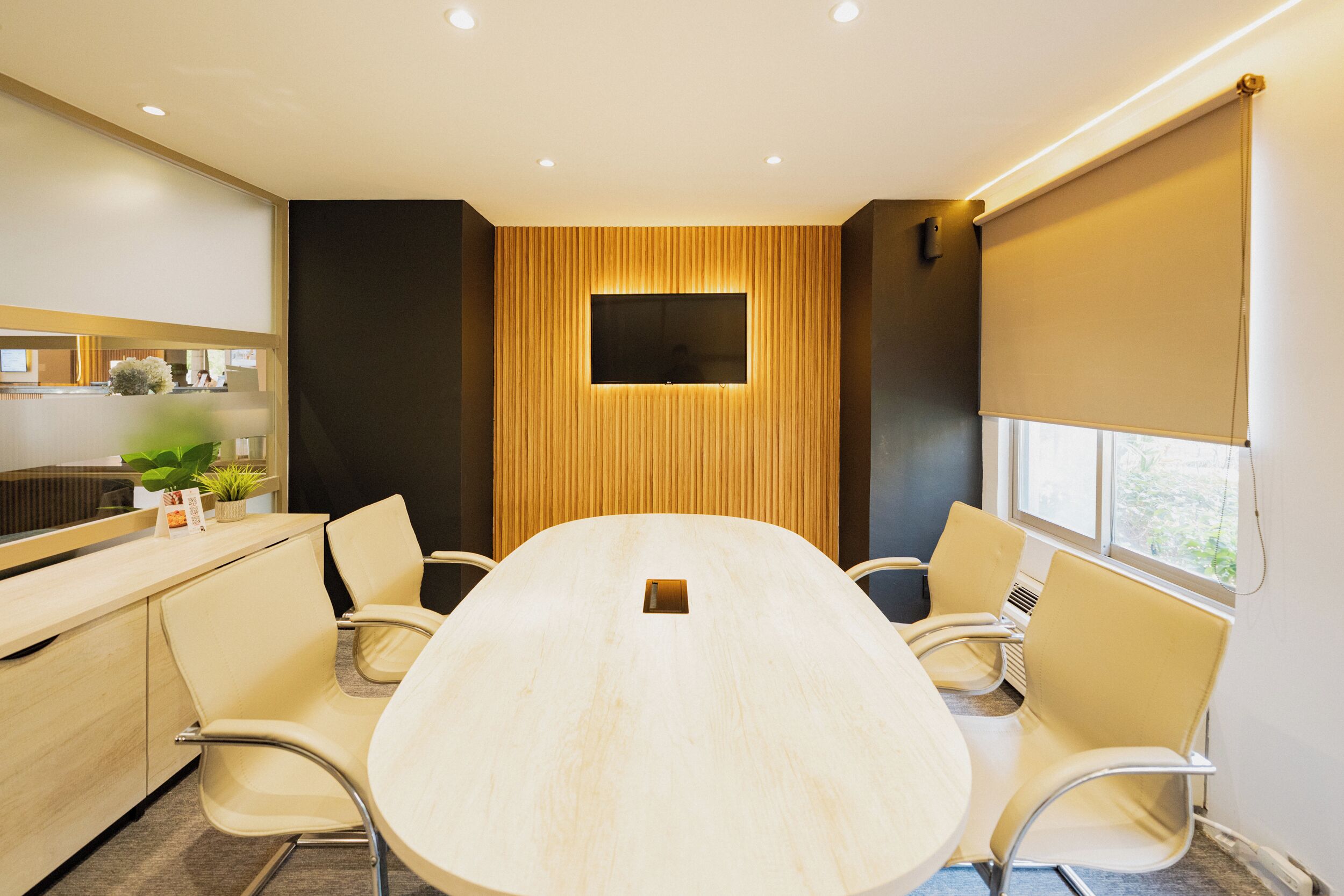 A conference room with a long table, six chairs, a TV screen, and a window with blinds.