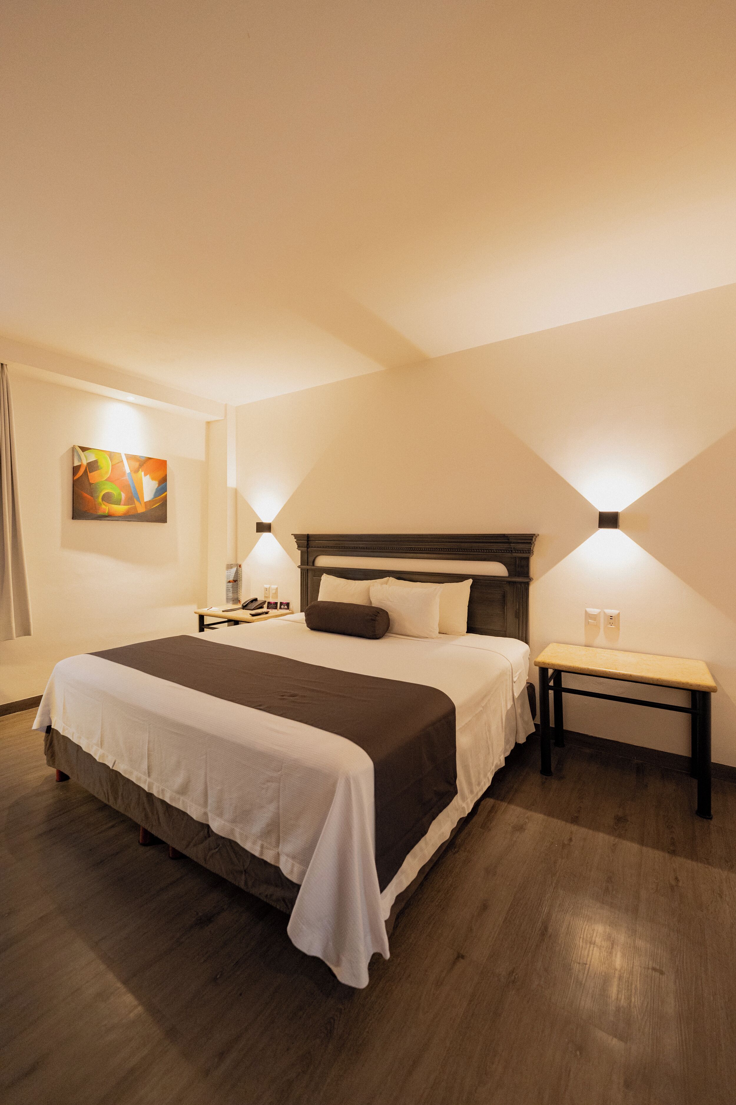 A hotel room with a large bed, bedside tables, a nightstand, a wall-mounted lamp, and a painting on the wall.