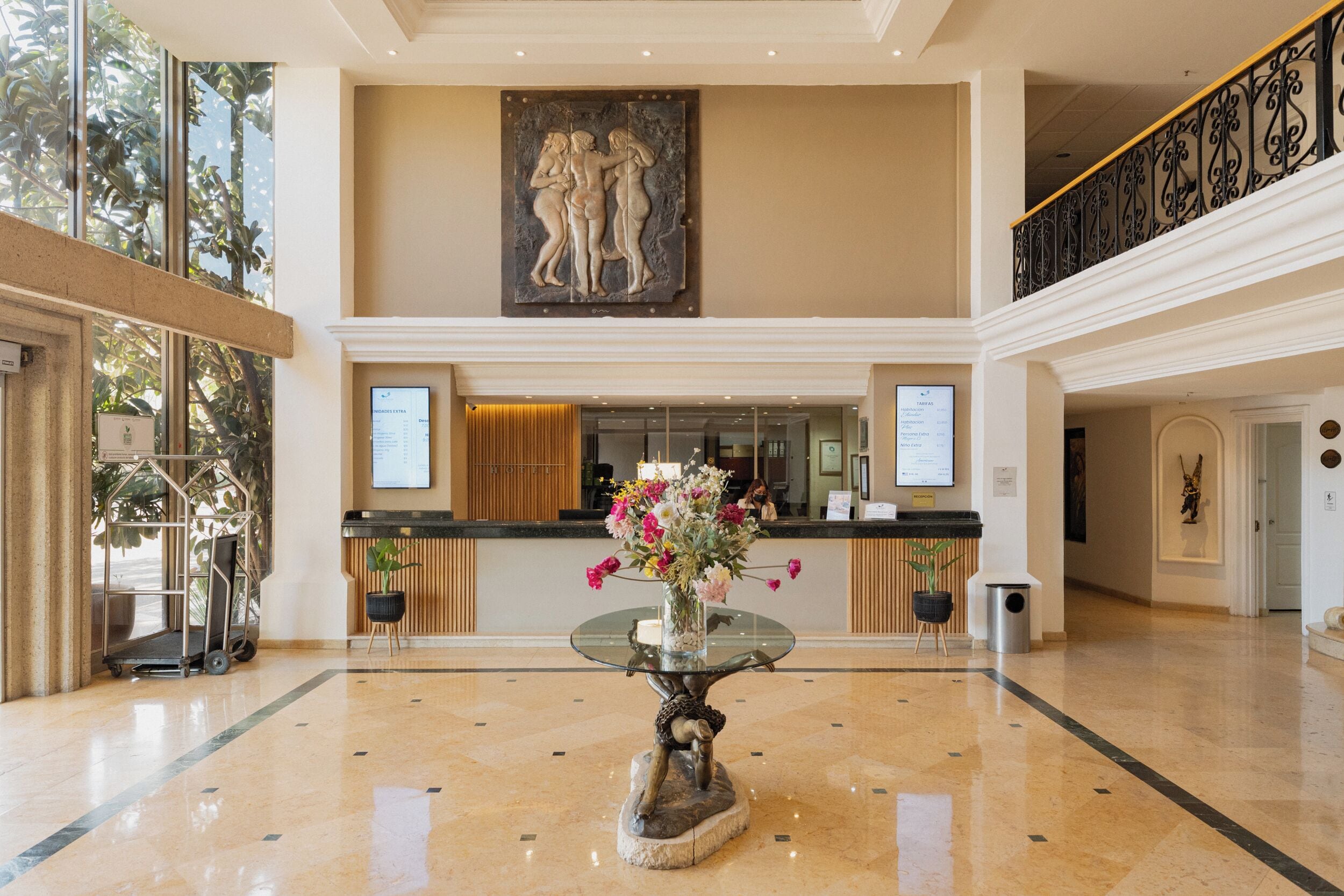 A lobby with a reception desk, a glass-top table with a vase of flowers, a large wall sculpture, and a staircase with a wrought iron railing.