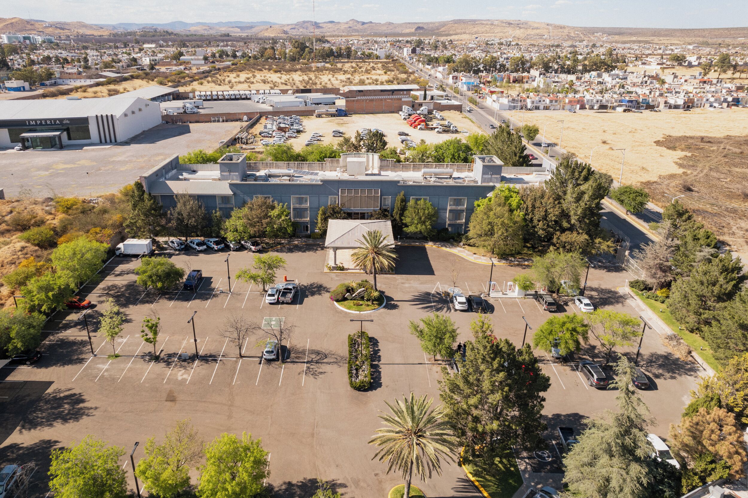 An industrial building with a parking lot, surrounded by trees and a residential area in the background.