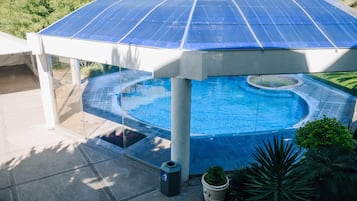 Een zwembad met een dak van zonnepanelen, omgeven door groen en een betonnen terras.