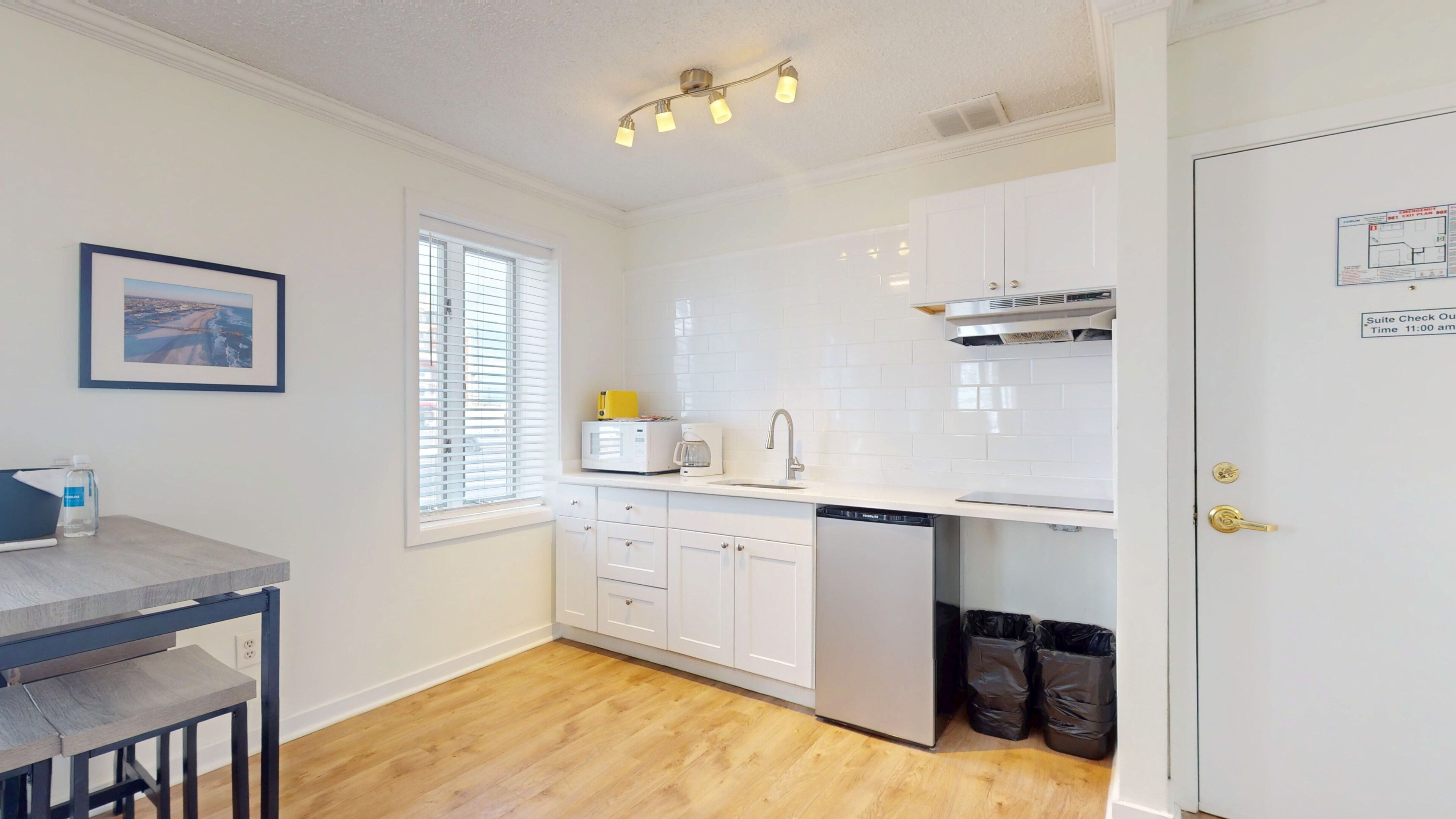A modern kitchen with white cabinets, a microwave, a coffee maker, and a dishwasher. There is a window with blinds, a framed picture on the wall, and a door with a keyhole.
