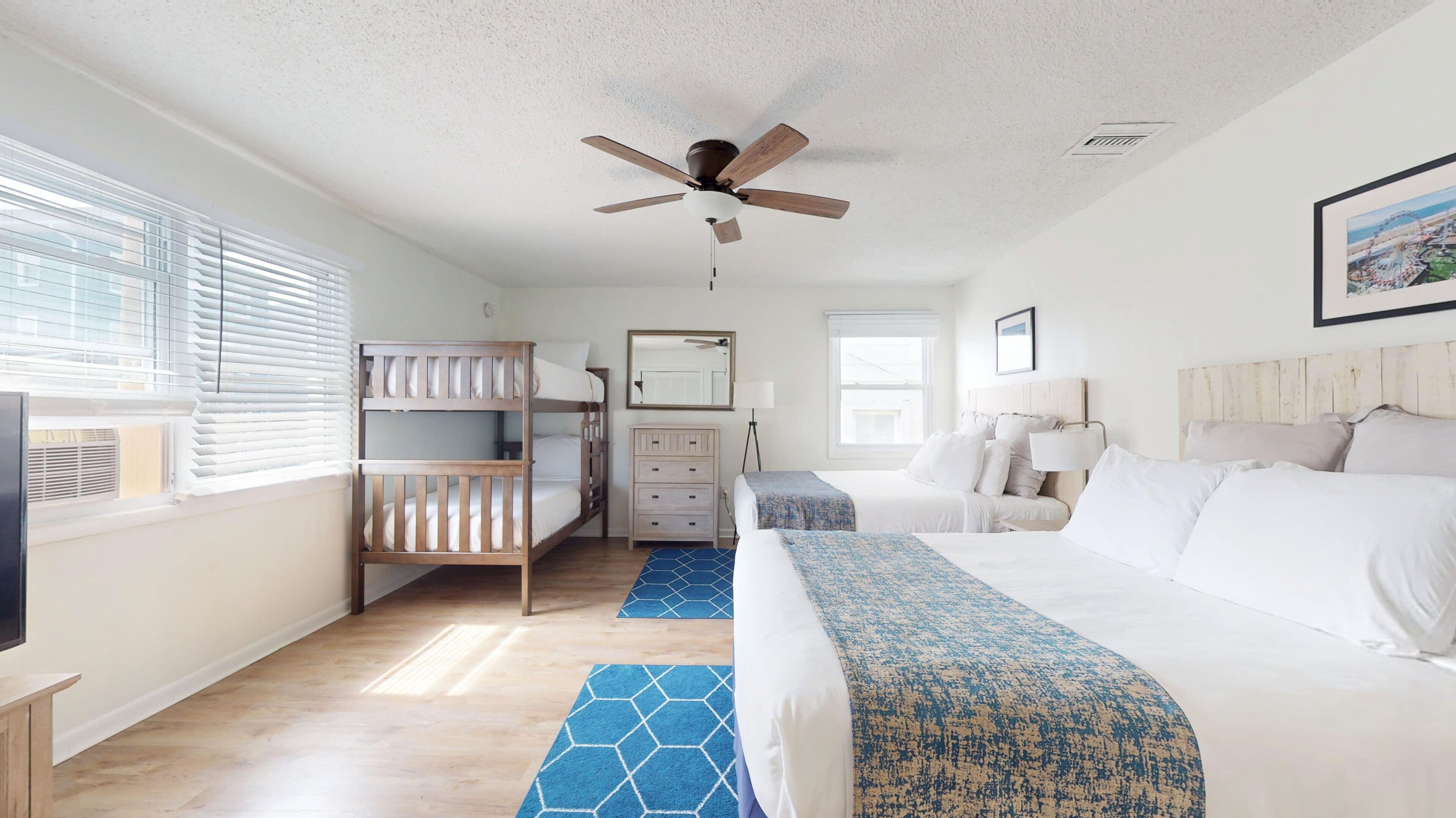 A hotel room with a bunk bed, a single bed, a ceiling fan, a desk, and a window with blinds.