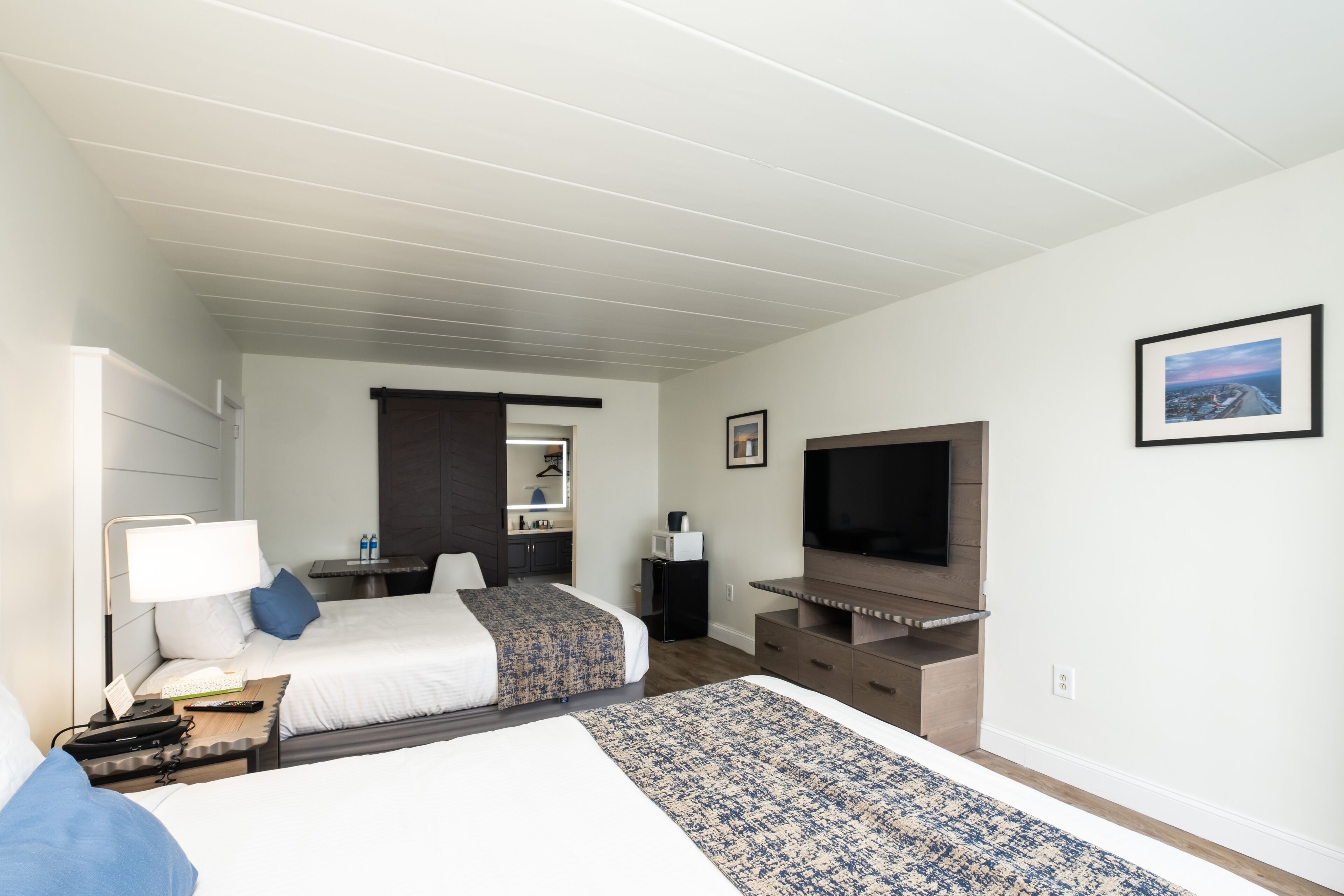 Quarto de hotel com duas camas, televisão, secretária com luminária, mesa pequena e um quadro na parede.