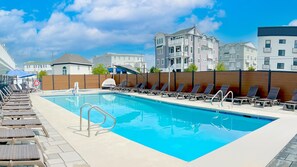 A swimming pool with lounge chairs and a lifebuoy, surrounded by a wooden fence and buildings in the background. - The Forum (Ocean City)