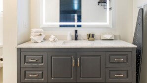 Un baño moderno con un mueble de tocador oscuro, un espejo grande con iluminación integrada y toallas ordenadas sobre el mueble.