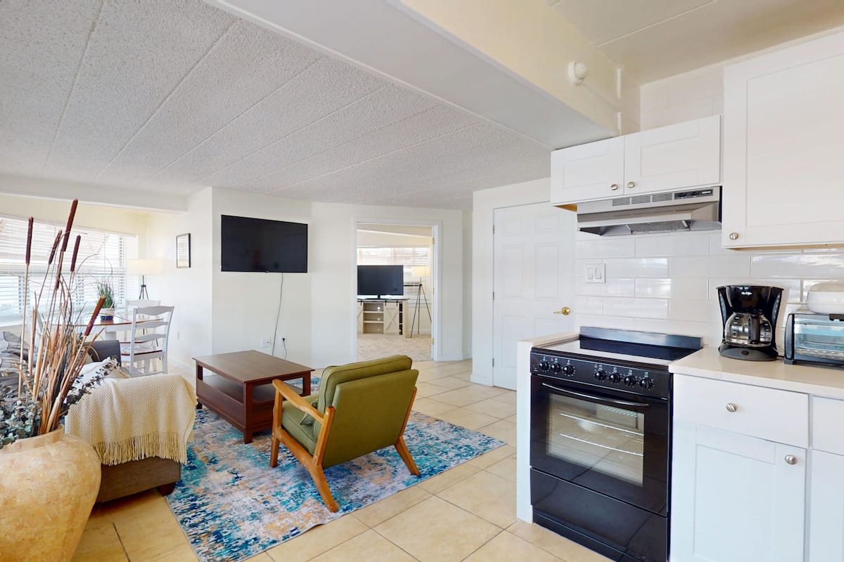 A modern kitchen with a stove, oven, and coffee maker, a dining area with a table and chairs, and a living room with a sofa and decorative plants.