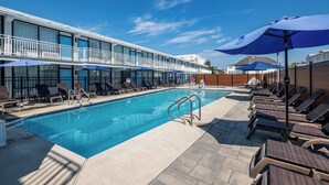Un área de piscina con sillones de descanso y sombrillas, una piscina y un edificio con balcones.